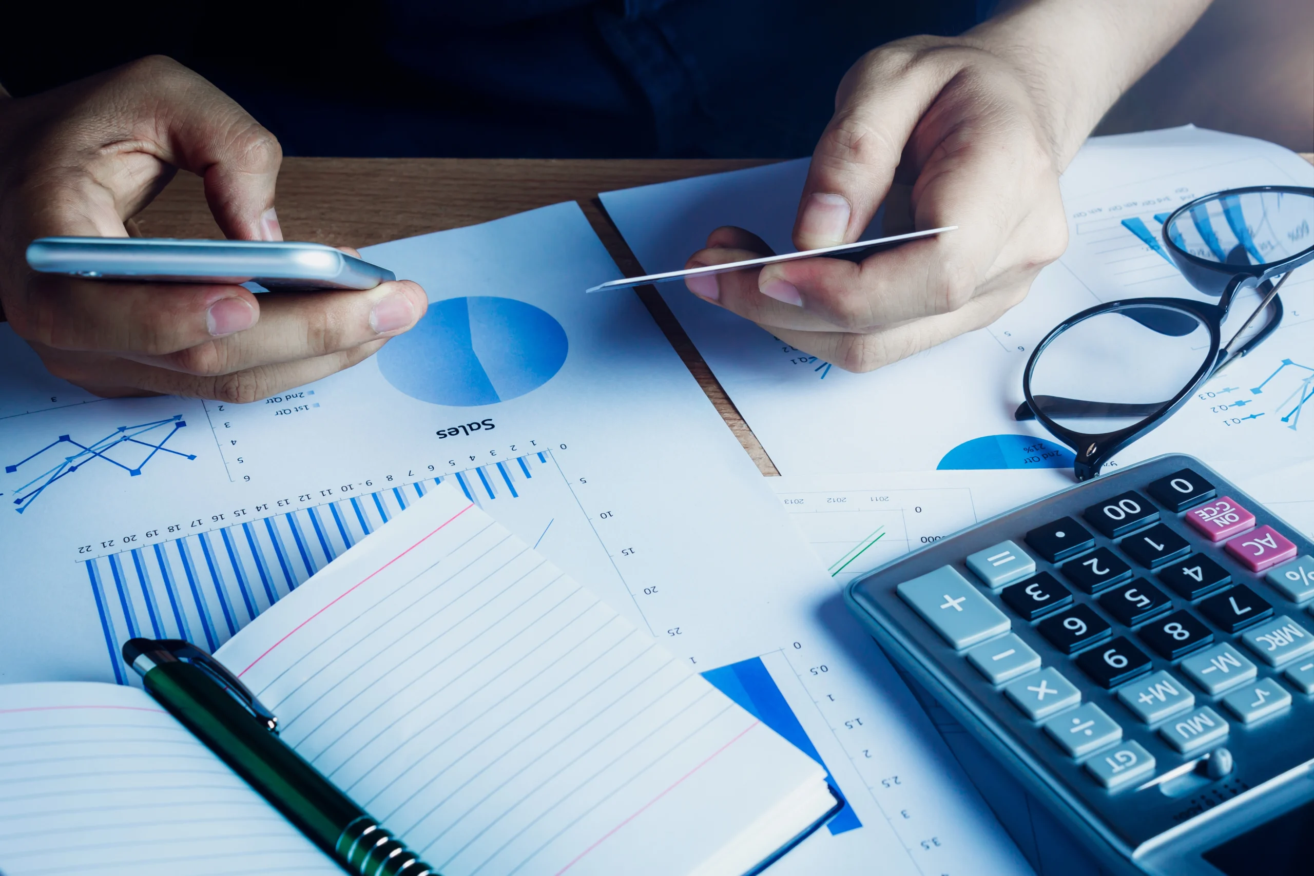 Un homme analyse des documents financiers liés aux fiches de paie, tenant une carte bleue et un téléphone, symbolisant la gestion des coûts et l'optimisation des dépenses salariales grâce à l’audit de fiche de paie.